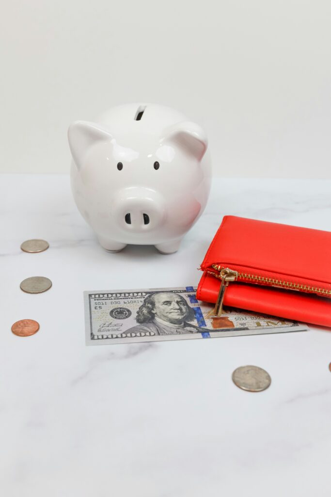 The best savings strategies for every income level A white piggy bank, scattered coins, a $100 bill, and a vibrant red wallet on a white marble surface, conveying themes of saving and finance.
